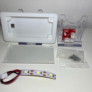 Puede incluir: Una carcasa de plástico rectangular blanca con una tapa con bisagras, una base de plástico transparente y un soporte de plástico transparente que sostiene una placa de circuito roja. También se incluye una tira de luces LED y una pequeña bolsa de tornillos.