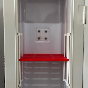 Puede incluir: Interior de un armario vintage abierto, de color blanquecino, con un estante rojo. El armario tiene forma rectangular con un acabado liso y brillante. El estante rojo está situado en el centro y el armario tiene un diseño sencillo y funcional.