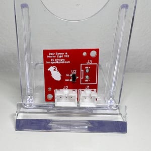 Puede incluir: Una placa de circuito roja con conectores blancos, etiquetada como "Door Sensor & Interior Light V1.2". El texto incluye "By Infragno" y una dirección de correo electrónico. Se encuentra en un soporte de plástico transparente.