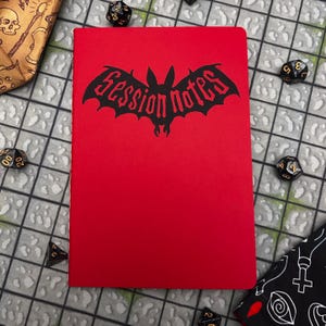 May include: A red notebook featuring the words "Session Notes" in a bat-shaped design. The notebook is surrounded by dice and a dice bag, suggesting a tabletop gaming theme. The cover is a solid, vibrant red.