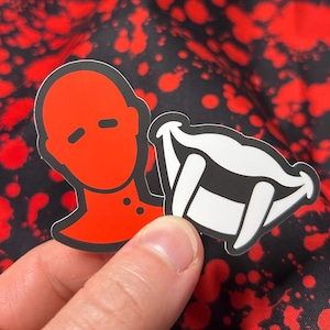 May include: Two stickers, one red and one white, depicting a faceless person and a set of fangs. The red sticker has a simple design of a person with a red face and black outline. The white sticker has a black outline and depicts a set of fangs.