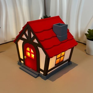 May include: A small, white and brown cottage with a red roof and a grey chimney. The cottage has a red door and two windows with yellow light shining through. The cottage is sitting on a brown surface.