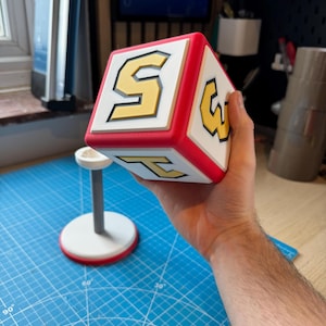 May include: A large cube in white, red, and yellow, displaying the numbers 5, 3, and 2 on its faces. The cube is held in a person's hand. A small stand in white and red is in the background.