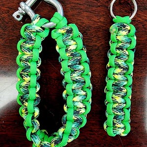 May include: Two handmade paracord accessories: a bracelet with a metal clasp and a keychain. Both are green with a camouflage pattern of yellow, black, and green. The bracelet is designed with a sturdy metal clasp.