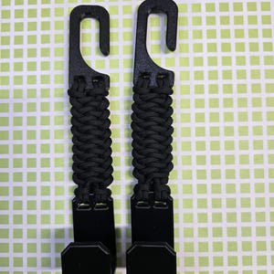 Puede incluir: Dos ganchos de pl&aacute;stico negro con un mango de paracord trenzado. Cada gancho tiene una secci&oacute;n superior curva para colgar y una secci&oacute;n inferior plana en forma de L. El cord&oacute;n negro est&aacute; tejido en un patr&oacute;n apretado entre los componentes.
