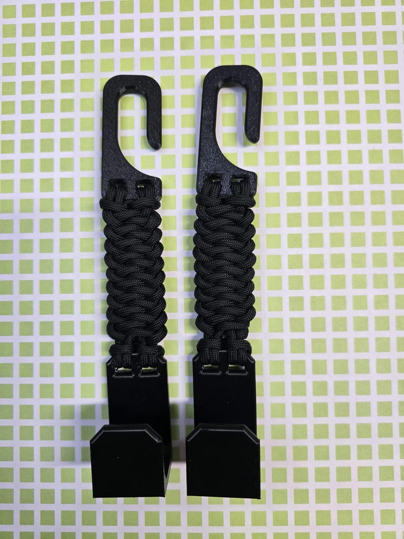 Puede incluir: Dos ganchos de pl&aacute;stico negro con un mango de paracord trenzado. Cada gancho tiene una secci&oacute;n superior curva para colgar y una secci&oacute;n inferior plana en forma de L. El cord&oacute;n negro est&aacute; tejido en un patr&oacute;n apretado entre los componentes.