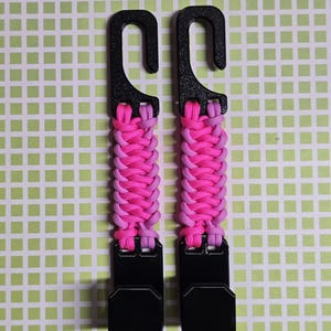 Puede incluir: Dos ganchos de pl&aacute;stico negro conectados por una correa de paracord trenzada en colores alternos rosa brillante y morado claro. Cada unidad cuenta con un gancho superior para colgar y un gancho inferior para asegurar objetos.