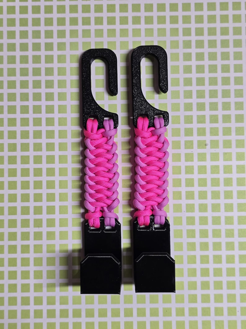 Puede incluir: Dos ganchos de pl&aacute;stico negro conectados por una correa de paracord trenzada en colores alternos rosa brillante y morado claro. Cada unidad cuenta con un gancho superior para colgar y un gancho inferior para asegurar objetos.