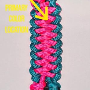 Puede incluir: Un gancho de pl&aacute;stico negro conectado a una correa de paracord trenzada en rosa ne&oacute;n y verde azulado, con una flecha amarilla que apunta a la secci&oacute;n rosa con el texto PRIMARY COLOR LOCATION, terminando en una base de pl&aacute;stico negro.