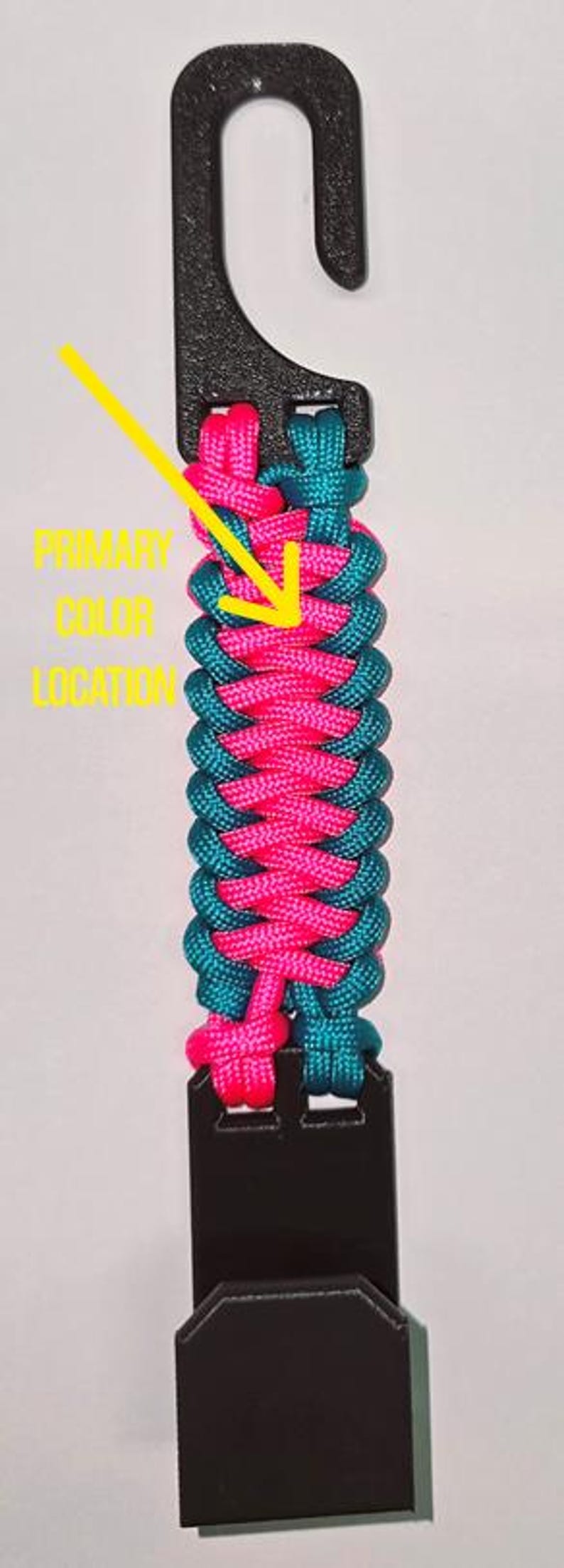 Puede incluir: Un gancho de pl&aacute;stico negro conectado a una correa de paracord trenzada en rosa ne&oacute;n y verde azulado, con una flecha amarilla que apunta a la secci&oacute;n rosa con el texto PRIMARY COLOR LOCATION, terminando en una base de pl&aacute;stico negro.