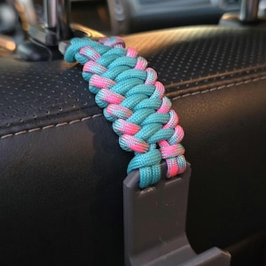 May include: A car seat headrest hook with a turquoise and pink braided paracord strap. The hook is gray and designed to attach to a car seat headrest for hanging items. The braided strap adds a decorative element.