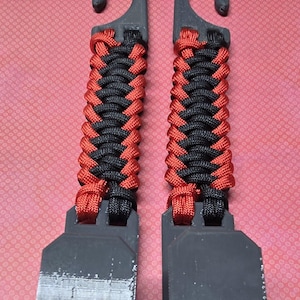 Puede incluir: Dos ganchos de pl&aacute;stico negro id&eacute;nticos con un mango de paracord trenzado en colores alternos rojo y negro. Cada gancho tiene una base plana rectangular y una secci&oacute;n superior curva, con el cord&oacute;n tejido firmemente entre los componentes.