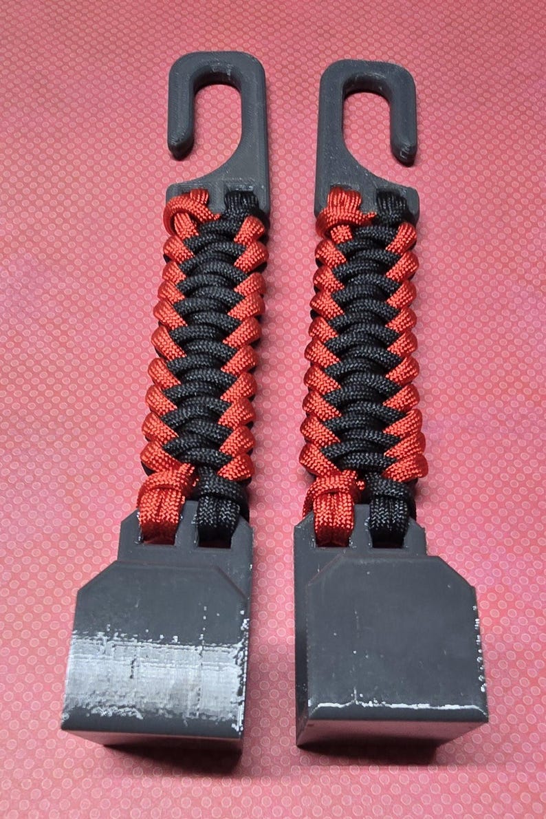 Puede incluir: Dos ganchos de pl&aacute;stico negro id&eacute;nticos con un mango de paracord trenzado en colores alternos rojo y negro. Cada gancho tiene una base plana rectangular y una secci&oacute;n superior curva, con el cord&oacute;n tejido firmemente entre los componentes.