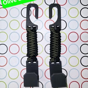 Puede incluir: Dos ganchos de pl&aacute;stico negro conectados por una correa de paracord trenzada en color oliva y negro. La parte superior de cada gancho tiene una abertura curva, mientras que la inferior tiene una base plana rectangular. Texto "Olive & Black".