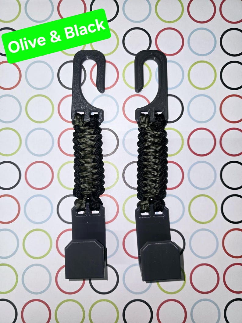 Puede incluir: Dos ganchos de pl&aacute;stico negro conectados por una correa de paracord trenzada en color oliva y negro. La parte superior de cada gancho tiene una abertura curva, mientras que la inferior tiene una base plana rectangular. Texto "Olive & Black".