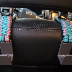 Puede incluir: Dos ganchos de pl&aacute;stico gris para el reposacabezas del coche con un envoltorio de paracord trenzado en colores alternos turquesa y rosa brillante. Los ganchos est&aacute;n sujetos a los postes met&aacute;licos de un reposacabezas de cuero negro.