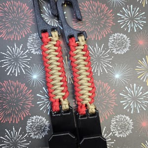 Puede incluir: Dos ganchos utilitarios de pl&aacute;stico negro conectados por una correa de paracord trenzada en rojo y beige. Cada correa cuenta con un gancho superior para colgar y un gancho inferior para asegurar objetos, con el cord&oacute;n tejido en un patr&oacute;n apretado.