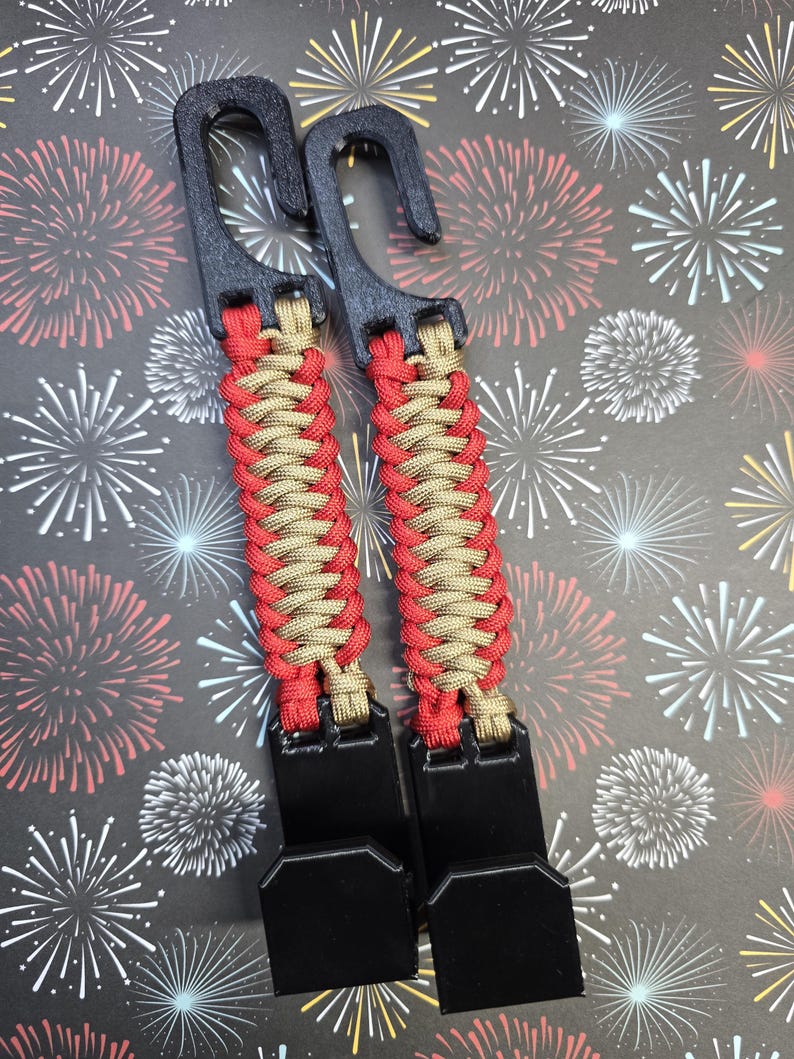 Puede incluir: Dos ganchos utilitarios de pl&aacute;stico negro conectados por una correa de paracord trenzada en rojo y beige. Cada correa cuenta con un gancho superior para colgar y un gancho inferior para asegurar objetos, con el cord&oacute;n tejido en un patr&oacute;n apretado.