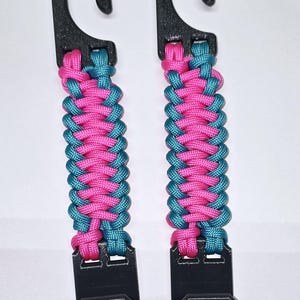Puede incluir: Dos ganchos para cochecito con herrajes de pl&aacute;stico negro conectados por una correa de paracord trenzada en colores alternos rosa brillante y azul cerceta. Cada unidad consta de un gancho superior y un clip inferior con textura.