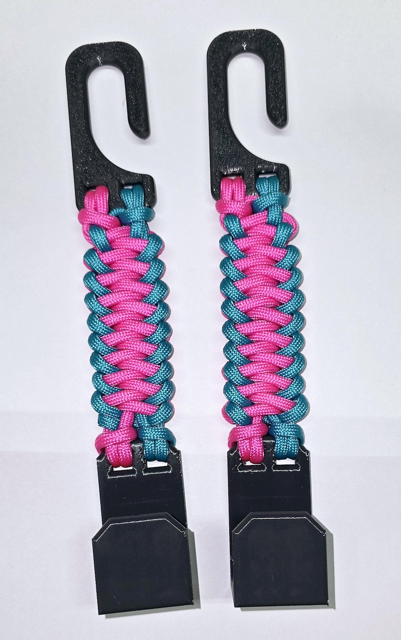 Puede incluir: Dos ganchos para cochecito con herrajes de pl&aacute;stico negro conectados por una correa de paracord trenzada en colores alternos rosa brillante y azul cerceta. Cada unidad consta de un gancho superior y un clip inferior con textura.