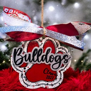 May include: A paw print shaped ornament featuring the word Bulldogs in black script and the text Class of 2027 in black. The ornament is red with white and black splatter accents and is topped with a multi-layered bow in red, white, and silver.