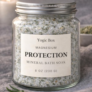 Puede incluir: Un tarro de vidrio transparente con tapa plateada lleno de sales de baño minerales. La etiqueta dice "Yogic Box MAGNESIUM PROTECTION MINERAL BATH SOAK 230 G". El tarro está sobre una superficie gris con una ramita de romero.