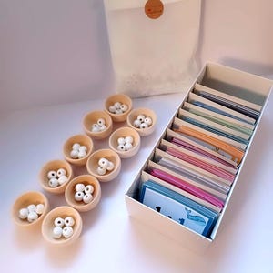 Puede incluir: Un conjunto de cuencos de madera con cuentas blancas, una bolsa de cuentas adicionales y una caja de tarjetas coloridas con números e imágenes de criaturas marinas.