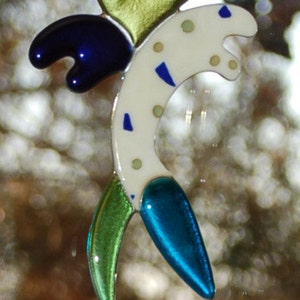 May include: A colorful glass art piece in the shape of a stylized fish. The fish features a white body with blue and green accents, a blue tail, and a green fin. A gold heart shape is at the top, and a dark blue shape is on the side.