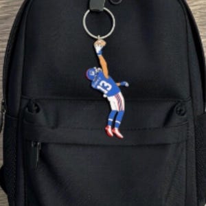 May include: A black backpack featuring a silver metal keyring attached to the top zip. The keyring holds a flat, colourful charm depicting a football player in a blue jersey with the number 13, captured in a mid-air jumping catch motion.