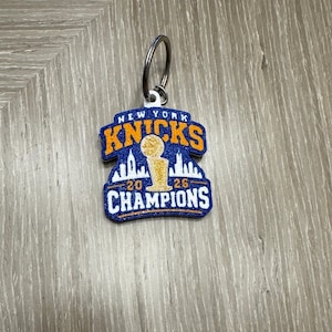 May include: A blue and orange glitter keychain featuring the text NEW YORK KNICKS at the top and CHAMPIONS at the bottom. The center displays a gold basketball trophy silhouette against a white skyline, with the numbers 20 and 26 on either side.