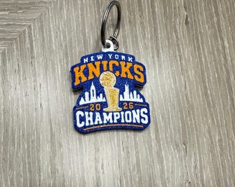 May include: A blue and orange glitter keychain featuring the text NEW YORK KNICKS at the top and CHAMPIONS at the bottom. The center displays a gold basketball trophy silhouette against a white skyline, with the numbers 20 and 26 on either side.