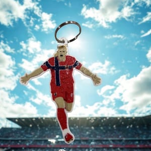 May include: A glittery acrylic keychain featuring a soccer player in a red jersey with a blue and white cross emblem, arms outstretched in a celebratory pose, attached to a circular metal split ring for keys or bag attachment.