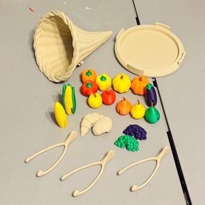 May include: A beige cornucopia with a matching circular lid, accompanied by miniature plastic fruits and vegetables including corn, pumpkins, apples, pears, eggplant, grapes, and three beige wishbones, all rendered in solid colors.