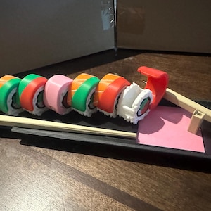 May include: A set of colorful sushi rolls on a black tray with chopsticks. The rolls are made with rice, seaweed, and various fillings. The tray is made of black plastic and has a pink mat on the side.