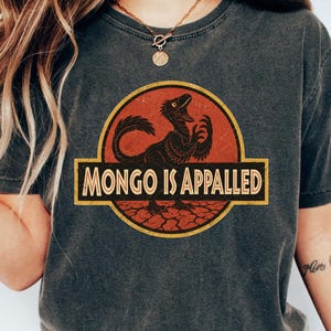 Könnte beinhalten: Dunkelgraues T-Shirt mit einem Dinosaurier-Motiv und dem Text "MONGO IS APPALLED" in Gelb. Die Grafik ist vor einem roten und orangefarbenen Hintergrund platziert. Ein lässiges Kleidungsstück.