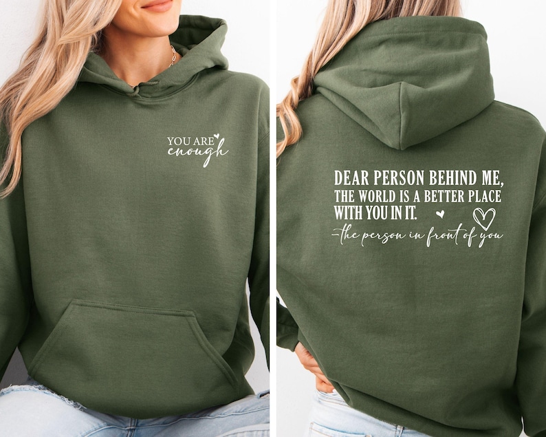 May include: An olive green hoodie with a front pocket and a hood. The front of the hoodie has the text "YOU ARE enough" in white script. The back of the hoodie has the text "DEAR PERSON BEHIND ME, THE WORLD IS A BETTER PLACE WITH YOU IN IT. the person in front of you."