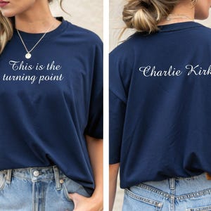 May include: Navy blue t-shirt with the text "This is the turning point" on the front and "Charlie Kirk" on the back. The shirt is a solid color and has a classic crew neck design. The shirt is paired with blue jeans.