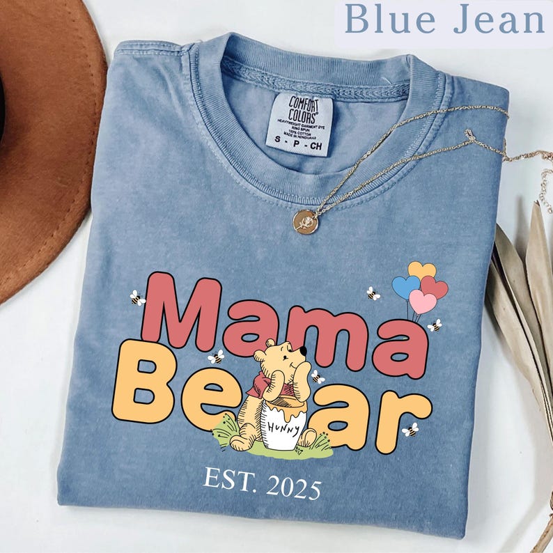 K&ouml;nnte beinhalten: Ein blaues Jeans-T-Shirt mit einem Winnie-the-Pooh-inspirierten Design. Das Shirt zeigt den Text "Mama Bear" in bunten Buchstaben mit einem Cartoon-Winnie-the-Pooh, der einen Honigtopf h&auml;lt. Der Text "EST. 2025" ist unter dem Design gedruckt.