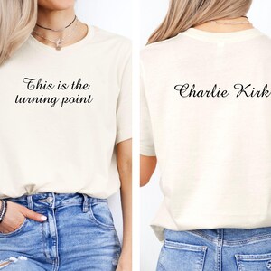 May include: Cream-colored t-shirt with the text "This is the turning point" on the front and "Charlie Kirk" on the back. The shirt is paired with blue jeans. The t-shirt has short sleeves and a crew neck.