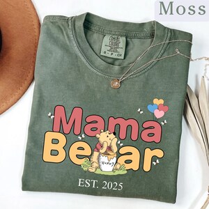 K&ouml;nnte beinhalten: Ein gr&uuml;nes T-Shirt mit einem Cartoon-Winnie-the-Pooh-B&auml;ren, der ein Honigglas h&auml;lt. Der Text "Mama Bear" ist in gro&szlig;en, bunten Buchstaben &uuml;ber dem B&auml;ren. Der Text "EST. 2025" befindet sich unter dem B&auml;ren.