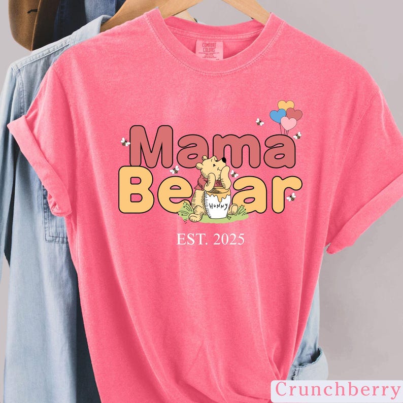 K&ouml;nnte beinhalten: Pinkes T-Shirt mit einem Comic-B&auml;ren, der einen Honigtopf h&auml;lt, und dem Text "Mama Bear EST. 2025".