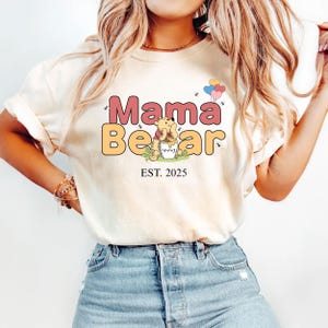 K&ouml;nnte beinhalten: Wei&szlig;es T-Shirt mit einem Aufdruck von Winnie Puuh, der einen Honigtopf und Ballons h&auml;lt. Der Text "Mama Bear" ist in rosa und gelben Buchstaben mit dem Jahr "EST. 2025" darunter.