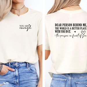 May include: Cream-colored t-shirt with black text on the front that reads "YOU ARE enough". The back of the shirt has the text "DEAR PERSON BEHIND ME, THE WORLD IS A BETTER PLACE WITH YOU IN IT." with a heart symbol and the text "-the person in front of you."