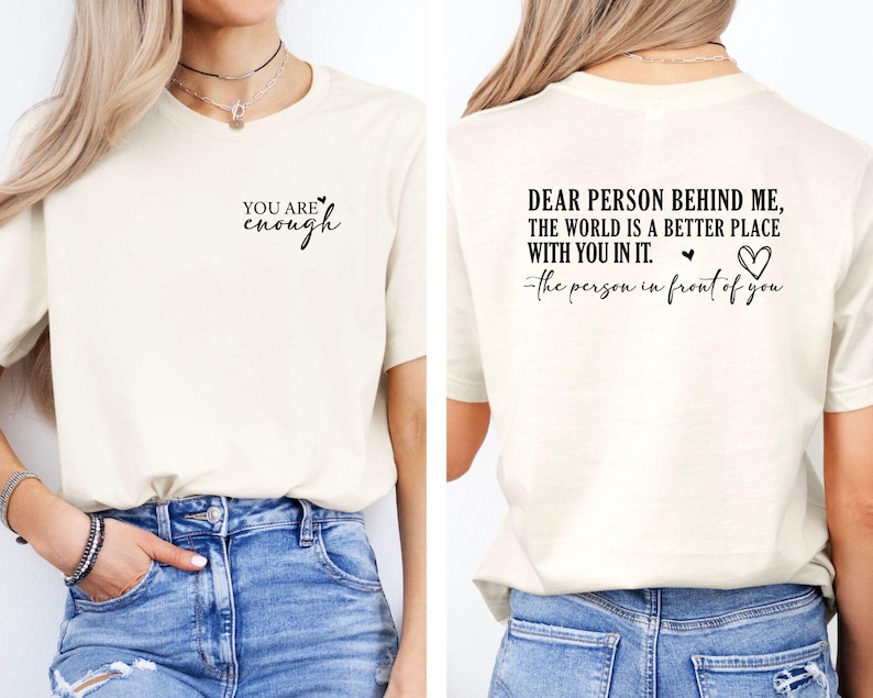 May include: Cream-colored t-shirt with black text on the front that reads "YOU ARE enough". The back of the shirt has the text "DEAR PERSON BEHIND ME, THE WORLD IS A BETTER PLACE WITH YOU IN IT." with a heart symbol and the text "-the person in front of you."