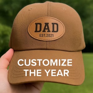 May include: Brown baseball cap with a leather patch that reads "DAD EST.2021". The text "CUSTOMIZE THE YEAR" is printed on the front of the cap. The cap is held by a hand.