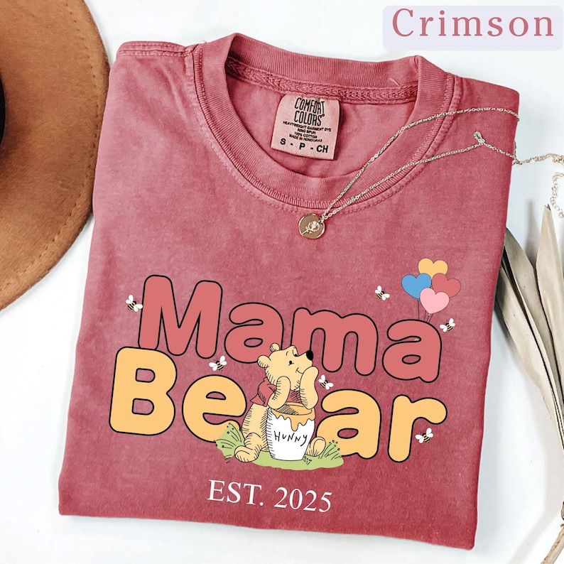May include: Crimson red t-shirt with a vintage-style graphic of Winnie the Pooh as a bear holding a pot of honey. The text "Mama Bear" is in large, colorful letters with the year "EST. 2025" below.