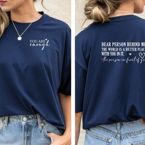May include: Navy blue t-shirt with the text "YOU ARE enough" on the front. The back of the shirt features the message "DEAR PERSON BEHIND ME, THE WORLD IS A BETTER PLACE WITH YOU IN IT. -the person in front of you."