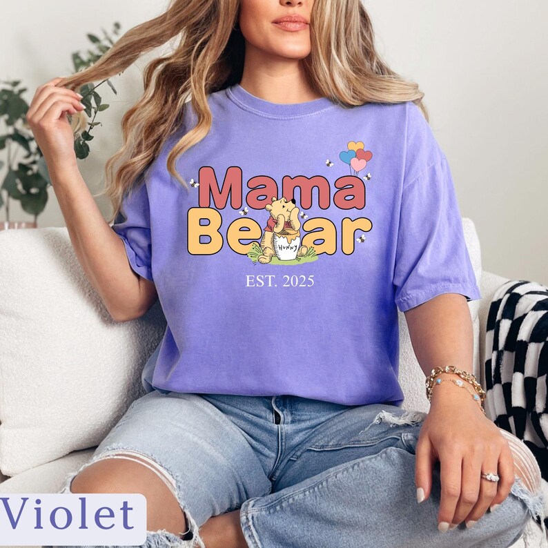K&ouml;nnte beinhalten: Lilafarbenes T-Shirt mit einem Aufdruck von Winnie Puuh, der einen Honigtopf h&auml;lt. Der Text "Mama Bear Est. 2025" ist auf dem T-Shirt aufgedruckt.