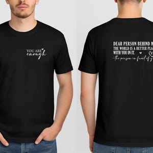 May include: Black t-shirt with white text on the front that reads "YOU ARE enough". The back of the shirt has the text "DEAR PERSON BEHIND ME, THE WORLD IS A BETTER PLACE WITH YOU IN IT. the person in front of you."