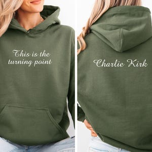 May include: Olive green hoodie with a front pocket and a hood. The front of the hoodie has the text "This is the turning point" in white script. The back of the hoodie has the name "Charlie Kirk" in white script.